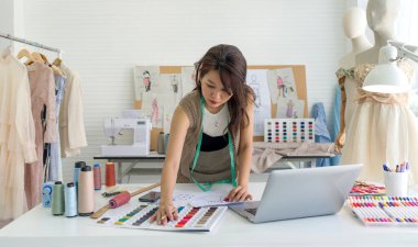 Bir kadın işine dalmış, yaratıcı materyallerle dolu parlak bir moda stüdyosunda dizüstü bilgisayarıyla çalışıyor..