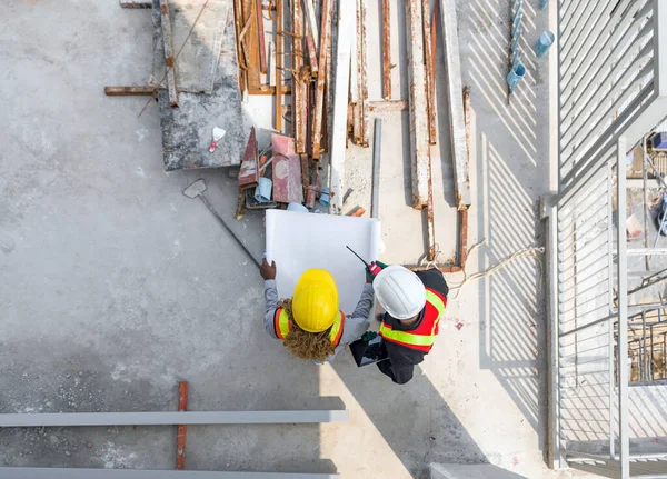 Two worker in helmet are busy, looking at blueprint on a sunny day at a construction site. High angle view.