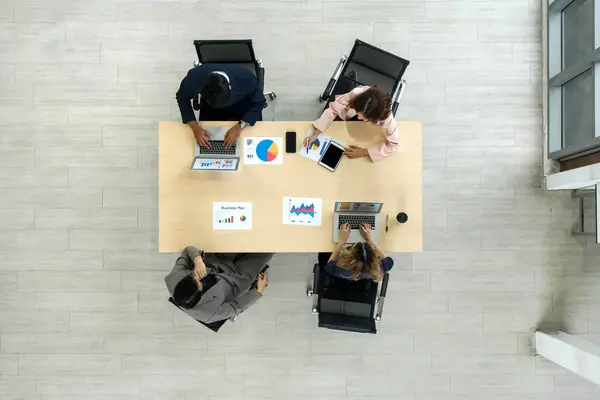 A team of workers is seated together at a big table in an office, sharing thoughts and talking. Top view