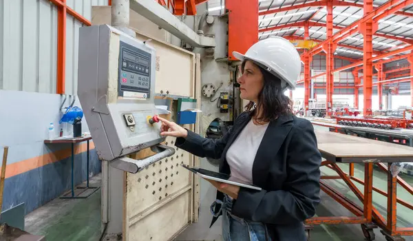 A woman in a hardhat is carefully observing a machinery monitor in a bustling factory.