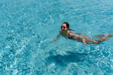 Bir kadın berrak, pırıl pırıl mavi bir havuzda süzülüyor, kolları rahatça yüzerken uzanıyor..