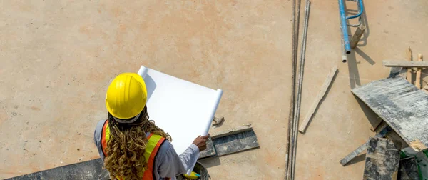 Worker in hardhat and reflective vest holding blueprint at a construction site on a sunny day. High angle view.