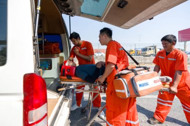 Turuncu üniformalı bir grup erkek hemşire, bir boyun ve kafa kazası kurbanını ambulansla bir yatağa yerleştiriyor..