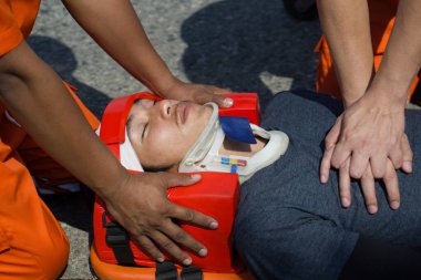 Uzun omurgalı sedyede yatıp emniyet kemeri takan baş sabitleyici aletli bir adam kurtarma ekibinden kalp masajı alıyor. Trafik kazasında acil yardım..