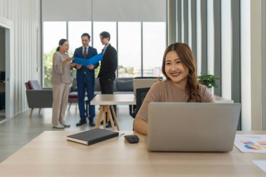 Genç bir kadın ön planda oturuyor, gülümsüyor ve dizüstü bilgisayarla çalışıyor. Arka planda, üç kişi daha bir sohbete katılıyor, bir belgeyi paylaşıyorlar. Ofiste geniş pencereli atmosfer.