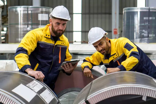 Two factory workers are inspecting a stack of large steel coils in an industrial setting or manufacturing facility.