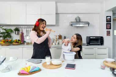 Mutfakta harika vakit geçiren iki kadın. Kulaklığı ve önlüğünün altında süveteri olan genç Asyalı bir kadın, elinde ahşap bir spatulayla şarkı söylüyor. Arkadaşı müziği dinlerken gülümsüyor..