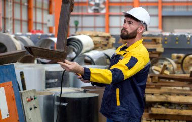 Kasket takan genç bir teknisyen ve güvenlik renginde yüksek görüş mesafesi olan bir ceket giyiyordu. Endüstriyel fabrikada makineleri denetliyordu. Üretim hattında makineler olan yoğun bir endüstri ortamı.