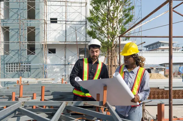 Two construction worker looking at a floor plan, standing in front of a large building site with helmet and safety gear. Day time work safety check. Work environment at the site of housing projects.