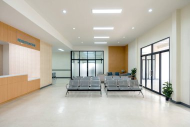 Hastanenin bekleme salonunda. Hasta ya da ziyaretçi randevu ya da hizmet için kayıt yaptırabilir. Bireysel bekleyiş için bağlı gri koltuklar var. Sağdaki cam kapı acil servise açılıyor..