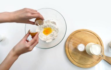 Yakından bir kase unun içine yumurta kırarak pişirmeye hazırlanıyor. Mutfak tezgahında kabartma tozu ve bir sürahi taze süt var. Üst görünüm