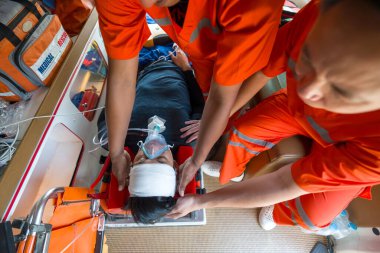 Oksijen maskesi takan biri ambulansın içinde turuncu üniformalı bir sağlık görevlisinden yardım alıyor. Üst görünüm
