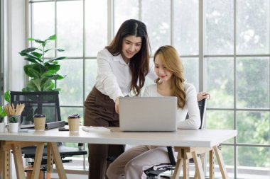 Two asian women working on laptop computer with a coffee cup, mobile phone and document folder at a desk. The background is a glass wall, overlooking the scenery of green nature
