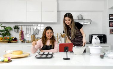 Two young women in an apron make dessert in the kitchen while looking at online recipe guide book on tablet computer.