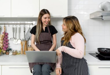 Two women with a striped apron reading or interacting with laptop computer screen in a modern kitchen. Cooking utensil and  decorated vegetable are on the countertop.