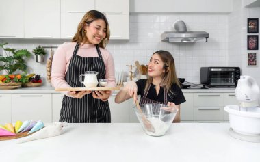 Two young women make dessert in the kitchen. One people stirring a white cake batter or icing mixture in a clear glass bowl. The other holding wooden tray with baking powder and a jug of fresh milk.