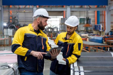 İş üniformalı ve koruyucu şapkalı iki fabrika işçisi endüstriyel bir ortamda veya imalat tesisinde büyük çelik bobinleri inceliyor..