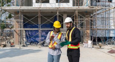 Two construction workers checking work schedule on tablet computer. Wear hardhat and safety vest, stands by metal scaffolding at a building site.