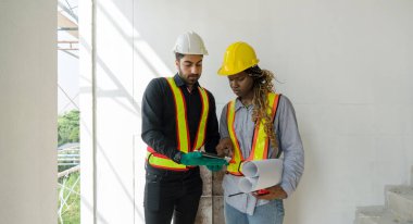 Tablet bilgisayardan çalışma saatlerini kontrol eden iki inşaat işçisi. Kask ve güvenlik yeleği giy, inşaat alanındaki merdivenlerin arasında dur..