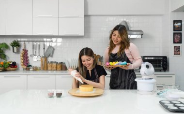 Önlüklü iki genç kadın mutfakta tatlı yapıyor. Bir kişi boru torbasıyla pastaya krema döker. Diğerinde bir avuç renkli fırın ya da dekorasyon malzemesi olan ahşap tepsi var..