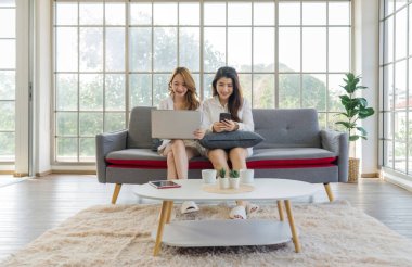 İki Asyalı kadın bir kanepede oturuyor, biri dizüstü bilgisayar kullanıyor ve diğeri cep telefonu kullanıyor, parlak bir odada..