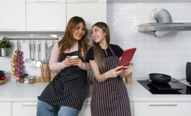 Çizgili önlüğü olan iki kadın modern bir mutfakta tablet bilgisayar ekranıyla etkileşime giriyor. Mutfak aletleri ve çeşitli sebzeler tezgahın üstünde..