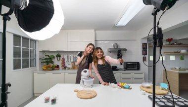 Önlüklü iki Asyalı kadın, mutfakta ellerinde akıllı bir telefonla gülümsüyor ve yemek pişiriyorlar. Selfie çubuğuyla video ve fotoğraf çekimi için ışıklandırma ekipmanları var. Sosyal blogcular için video kaydediyorlar..