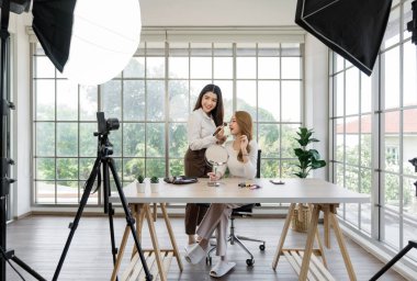 Sosyal medya için içerik yaratan iki Asyalı kadın. Tripoda monte edilmiş bir kamera, ışıklandırma ekipmanı ve masadaki çeşitli makyaj ürünlerini içeren bir stüdyo düzeneği kullanıyorlar..