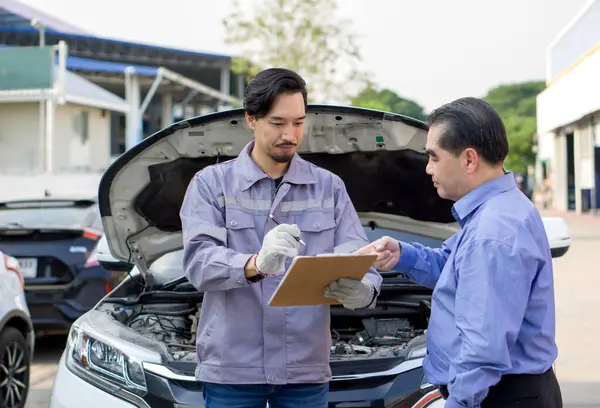 Eldivenli, iş üniformalı bir tamirci elinde evrak işlerine odaklanmış bir belge panosu tutuyor. Aracın sahibi ya da servis danışmanı aracın sorunlarını ya da bakım işlerini teknisyenle tartışıyor.