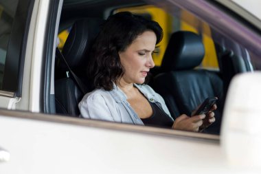 Bir kadın arabada oturuyor, telefonuna bakıyor, üzerinde önemli bir şey yazıyor ya da okuyor gibi görünüyor..