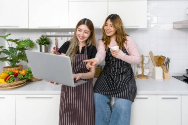 Çizgili önlüğü olan iki kadın modern bir mutfakta dizüstü bilgisayar ekranı okuyup etkileşimde bulunuyor. Mutfak aletleri ve çeşitli meyve ve sebzeler tezgahın üzerinde..