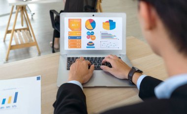 Back view of businessman in black suit sit at desk in office typing on laptop computer keyboard, making presentation with graph and multicolored chart.