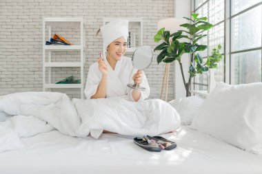 Gülümseyen Asyalı genç bir kadın, makyaj yapıyor, ayna tutuyor, kafasında bornoz ve havlu var..