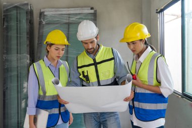 Miğfer ve güvenlik yeleği giymiş genç beyaz ustabaşı inşaat planına bak. Konut projesinin inşaat sahasında çalışma ortamı.