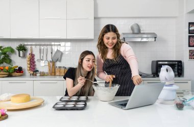 Önlüklü iki genç kadın, dizüstü bilgisayarda online tarif rehberine bakarken mutfakta tatlı yapıyorlar..