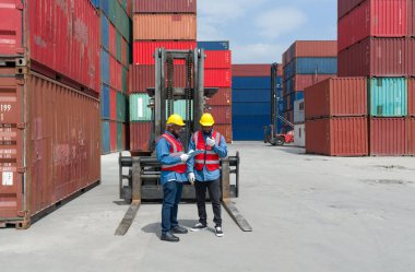 Kasketli, güvenlik yeleği ve koruyucu eldiven giymiş iki nakliye işçisi gün ışığında çalışıyor. Çalışma alanında ağır yük konteynırları var..
