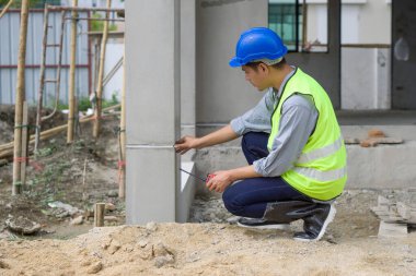 İnşaat miğferi ve güvenlik yeleği giymiş genç bir mühendis telsizi tutarken evin yapısını kontrol ediyor. Konut projelerinin inşaat sahasında mühendislerin çalışma ortamı