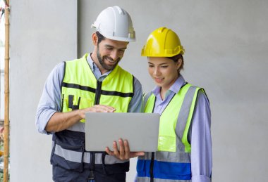 Dizüstü bilgisayarı olan genç bir mühendis ustabaşına kat planını açıklıyor. İkisi de miğfer ve çelik yelek giyiyor. Konut projelerinin inşaat sahasında çalışma ortamı.