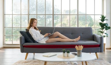 Beyaz gömlekli ve kısa pantolonlu genç Asyalı bir kadın, bir koltukta oturuyor, dizüstü bilgisayarda klavyeyle yazı yazıyor, geniş pencereli parlak bir odada rahatlıyor..