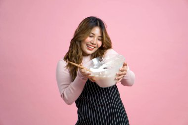 Önlüklü mutlu bir kadın elinde hamurlu ve tahta kaşıklı cam bir kâse tutuyor. Pembe arka planda stüdyo ışığıyla portre.