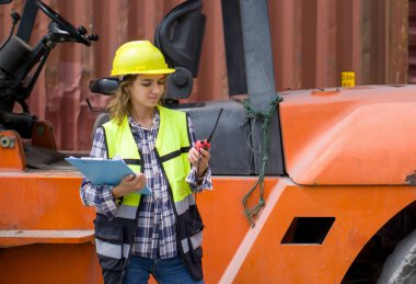 Sarı kasketli ve güvenlik yeleği olan beyaz bir kadın telsiz ve pano tutarken forklift önünde duruyor. Çalışma alanında konteynır var..
