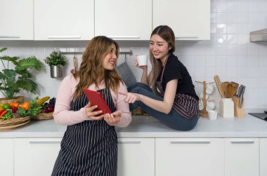 Two women with a striped apron reading or interacting with tablet computer screen in a modern kitchen. Cooking utensil and an assortment of decorated fruit and vegetable are on the countertop.