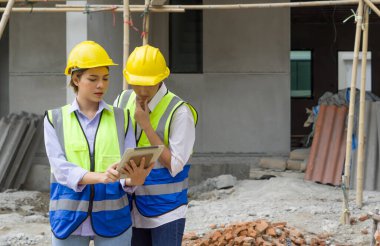 İnşaat miğferi ve güvenlik yeleği giymiş genç bir mühendis ustabaşına inşaat halindeki projeleri anlatıyor. Konut projelerinin inşaat sahasında mühendislerin çalışma ortamı