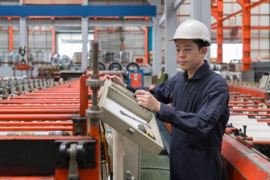 Kask ve mekanik giysisi olan bir adam, işlek bir fabrika ortamında endüstriyel ekipman kullanıyor ve makineleri ustalıkla yönetiyor..