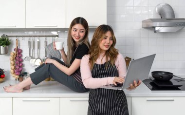 Çizgili önlüğü olan iki kadın modern bir mutfakta dizüstü bilgisayar ekranı okuyup etkileşimde bulunuyor. Mutfak aletleri tezgahın üstünde..