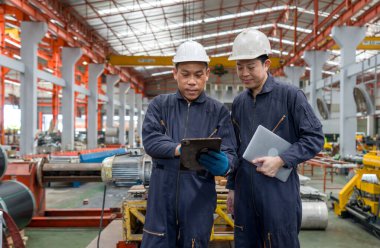 Güvenlik kaskı ve mekanik giysili iki işçi bir fabrikadaki tablete bakıyor. Bilgileri kontrol eder ve görevlerini tartışırlar..