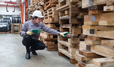 Baret ve koruyucu eldiven takmış bir işçi, bir fabrikada oturup evrak panosunu tutarken malzemeleri kontrol ediyor. Çevre, metal ve ahşap paletler ile düzenlenmiştir..