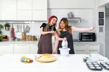 Mutfakta harika vakit geçiren iki kadın. Önlüğün altında Asyalı genç bir kadın, ahşap spatulayla şarkı söylüyor. Arkadaşı müziği dinlerken gülümsüyor..