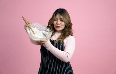 Önlük giymiş, hamuru tahta kaşıkla karıştıran mutlu bir kadın. Pembe arka planda stüdyo ışığıyla portre. 