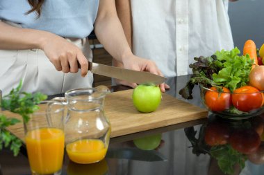 Yakından, tahta bir tahtanın üzerinde yeşil elma kesen bir bıçak tutan el. Karışık meyve suyu ve çeşitli meyve ve sebzelerle dolu cam kase mutfak tezgahına konur..